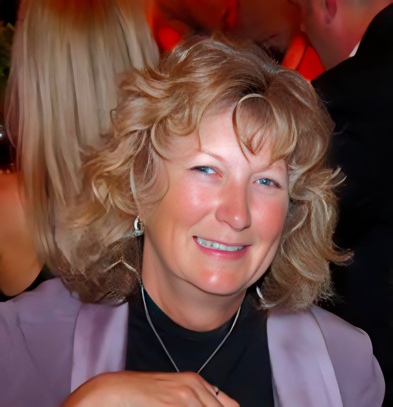 A woman with wavy blonde hair, wearing a lavender blazer and black top, smiles while seated indoors at a social event.