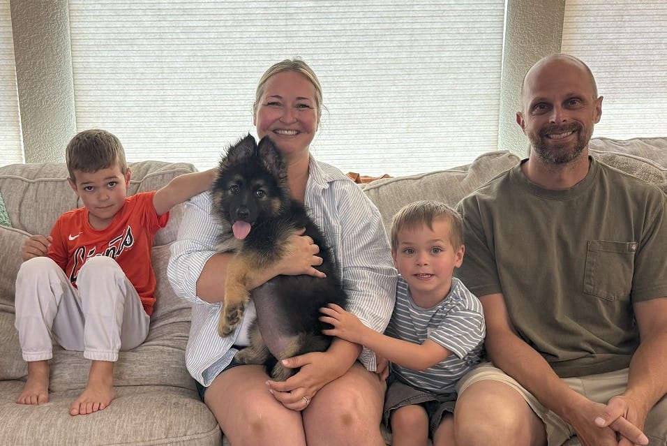 A family of four sits on a couch with a German Shepherd puppy; two boys are seated on either side of a smiling woman holding the dog, and a man sits at the end.